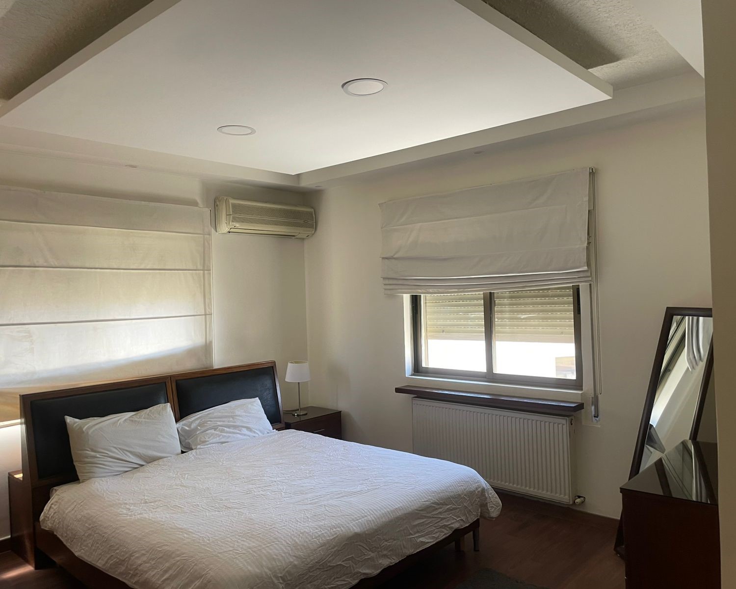 77600f4d-51ac-4113-97ce-a7da04bdd9d0 Bedroom with white quilted bed, two pillows, wooden headboard, and nightstand lamp on the left side.