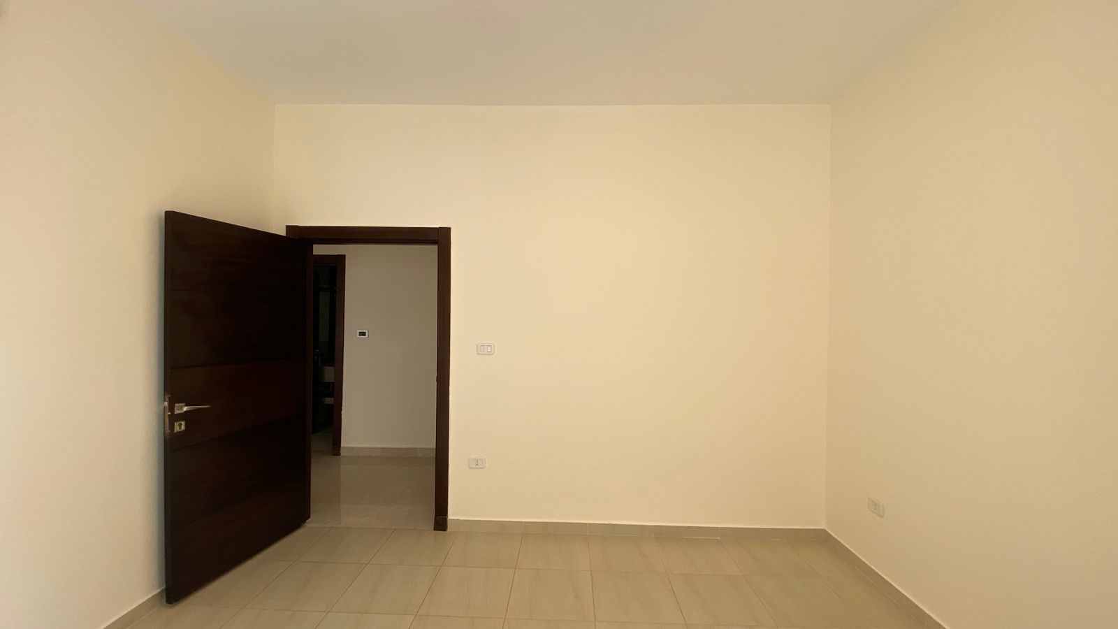 7797a6ce-0588-46bb-a5d3-1cc29fca867f Empty beige-walled room with a dark wooden door ajar, showing another hallway and doorway beyond.