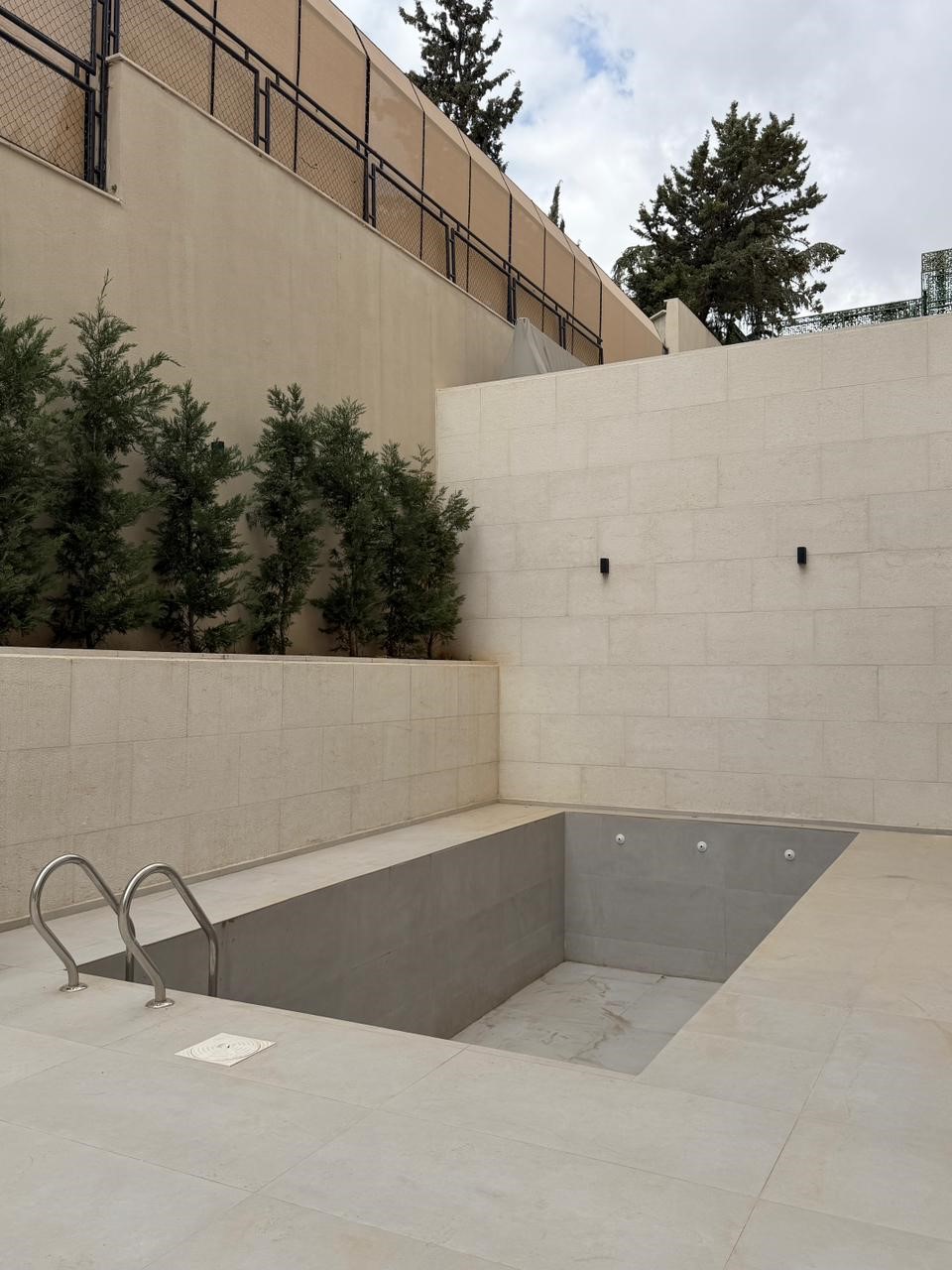 78aa3a4a-49e8-4e60-99de-3cda8711b39b Empty rectangular outdoor pool with a metal ladder, surrounded by beige stone walls and a row of evergreen shrubs.