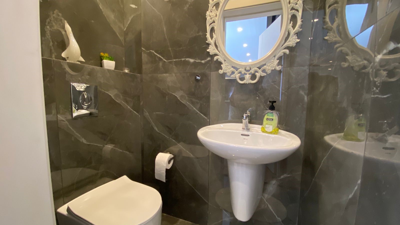 78c33f24-ebf0-4788-8aa4-36ab27eb7bc9 White pedestal sink with ornate round mirror above, hand soap on the rim, in a marble-tiled bathroom.