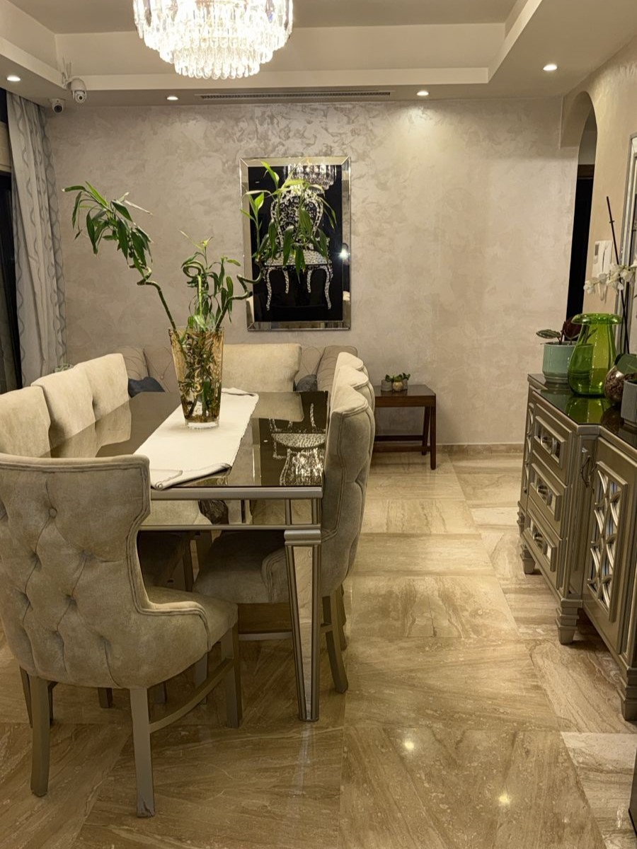 790b675d-63d1-4212-a60d-10d414928f22 Elegant dining room with beige upholstered chairs around a glass-topped table and a crystal chandelier above. A tall plant centerpiece sits in the middle of the table.