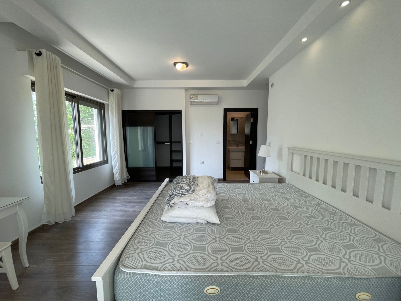 793fa8cc-7548-40be-8682-0d8721e0ad41 Bright hotel-style bedroom with a white wooden bed frame, patterned mattress, and a nightstand with a lamp on the right; large window with white curtains on the left and a wardrobe opposite the bed.