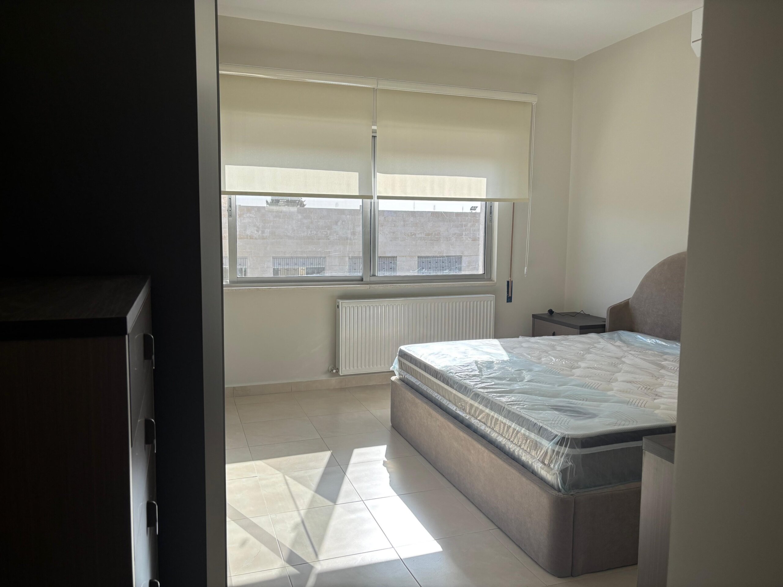 79b914bc-f345-43e7-b2aa-f1dcb3a8e81a Bright minimalist bedroom with a low upholstered bed, plastic-wrapped mattress, window with roller blinds, radiator beneath the window, and a small nightstand nearby.