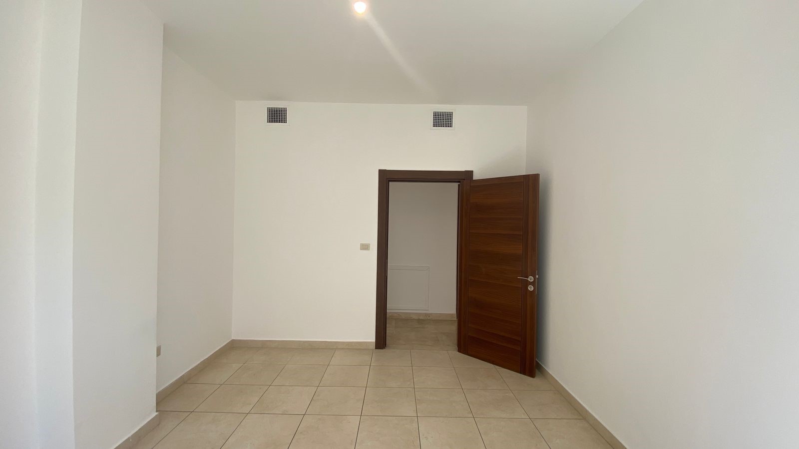 7a39f98b-e23e-4c9b-8f80-5743afc9f2ff Empty white room with beige tile floor and a dark wood door partly open leading to a small hall.