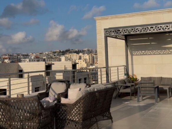Rooftop terrace with dark wicker seating, light cushions, and a seating area, overlooking a cityscape under a blue sky.