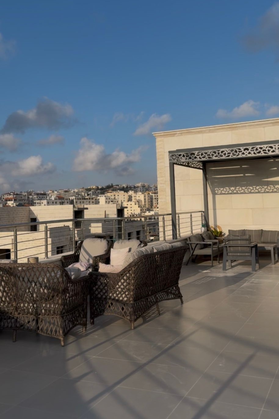 7acaf084-8b3a-4475-a4db-ca15c8ea9219 Rooftop terrace with dark wicker seating, light cushions, and a seating area, overlooking a cityscape under a blue sky.