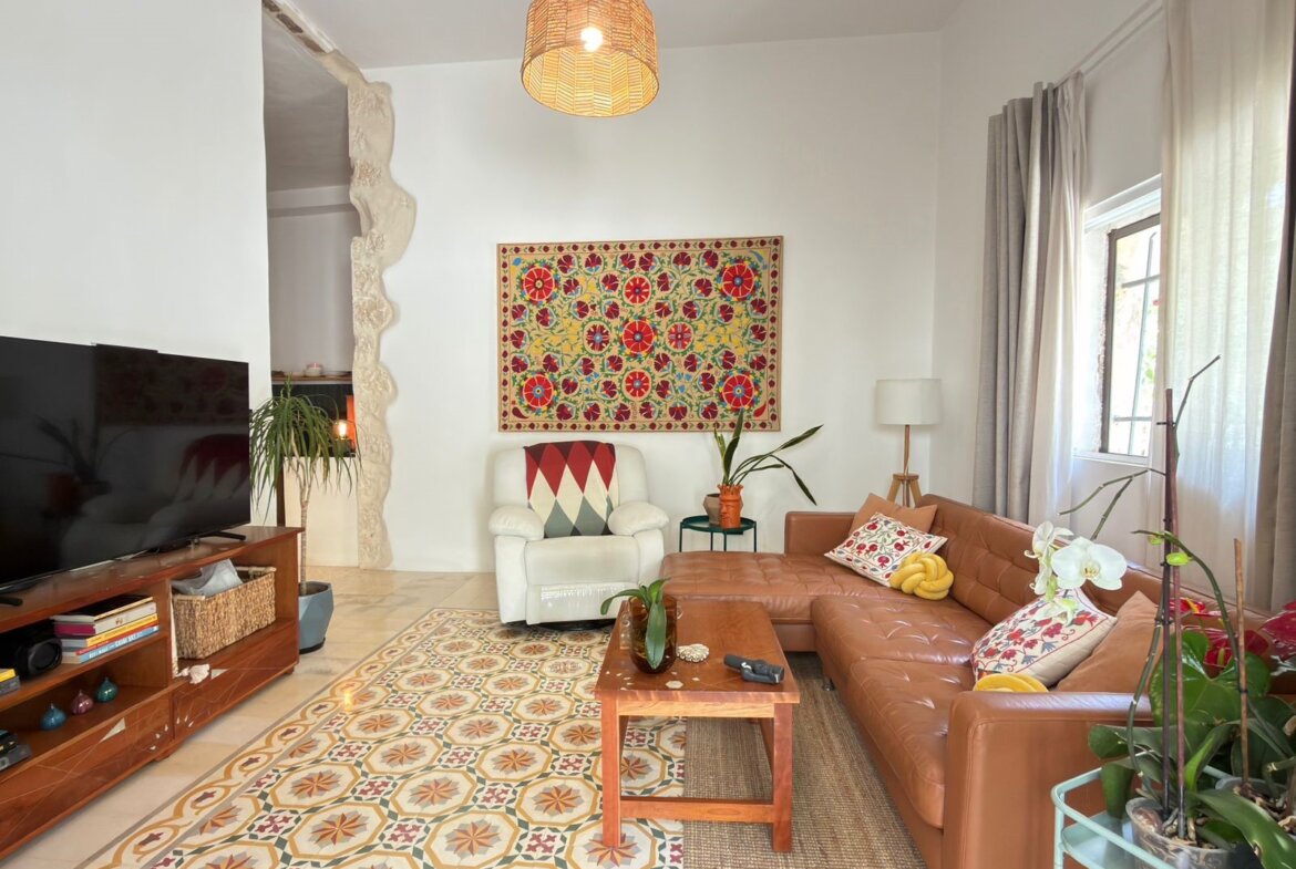 Living room with a brown leather sectional, a white armchair, and a large colorful wall hanging above the chair; windows with sheer curtains on the right, plants and a patterned rug on the floor.