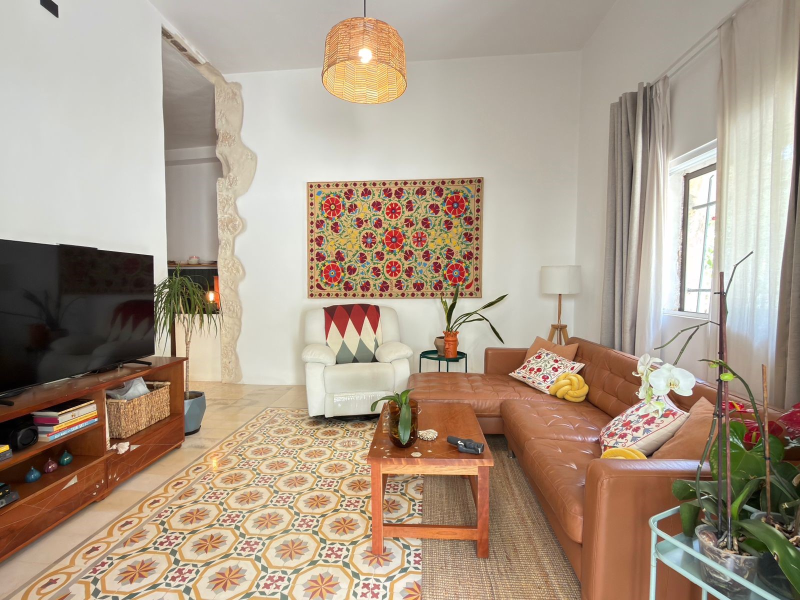 7b067edb-f316-4bb7-8f0f-0a95e9cb374c Living room with a brown leather sectional, a white armchair, and a large colorful wall hanging above the chair; windows with sheer curtains on the right, plants and a patterned rug on the floor.