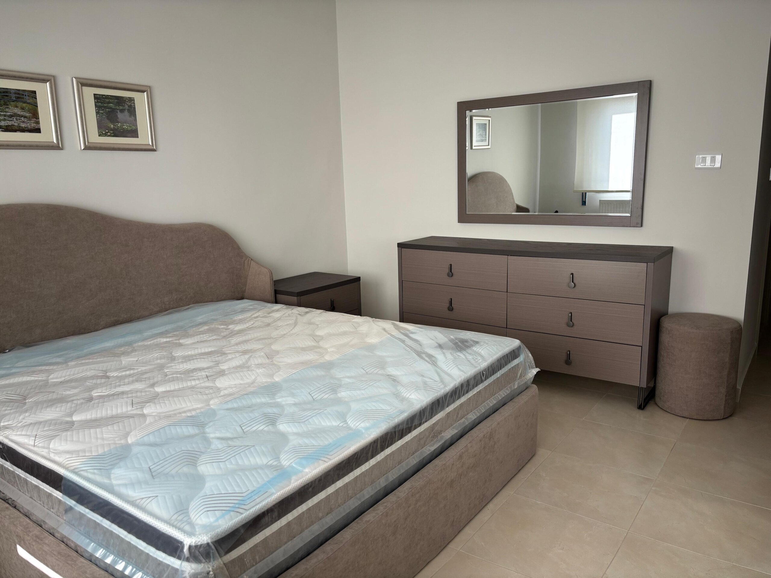 7b9c4d6b-f7fe-42c8-aad3-8abf7fbfc7d3 Queen bed with beige upholstered headboard, two matching nightstands, and a six-drawer dresser with a large framed mirror above. A round beige stool sits beside the dresser.