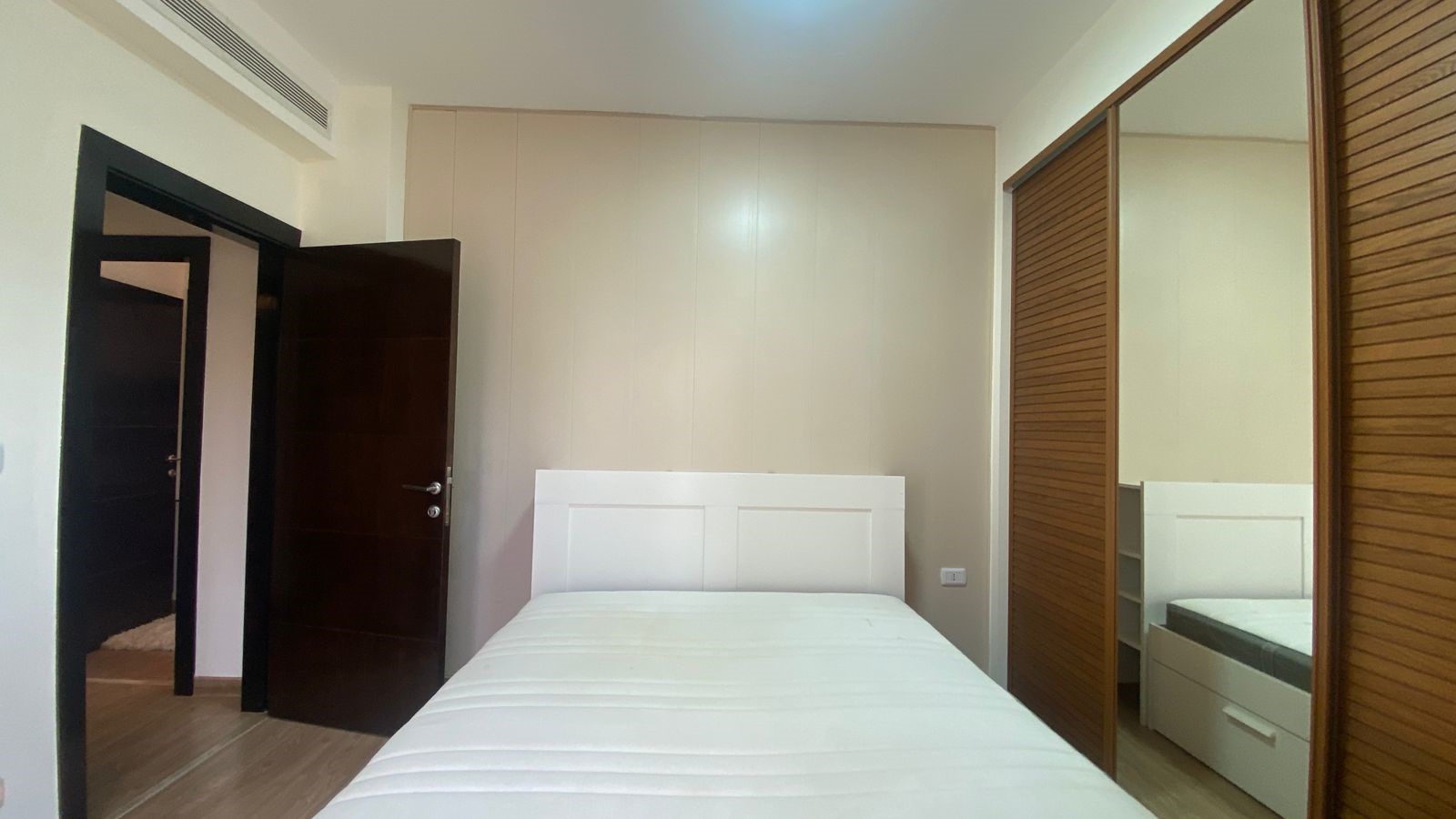 7c865a28-4065-4536-abaf-811d77c5cf5b Modern bedroom with white bed and headboard, beige walls, and large mirrored wardrobe doors to the right.