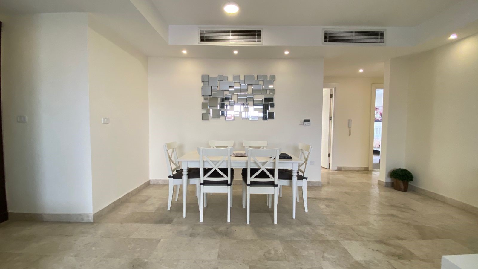 7d090ce9-210a-4cf4-9860-7d9db7277b93 White dining table with six white chairs in a bright, minimalist dining area; a mirror-square wall art piece is centered on the wall behind the table