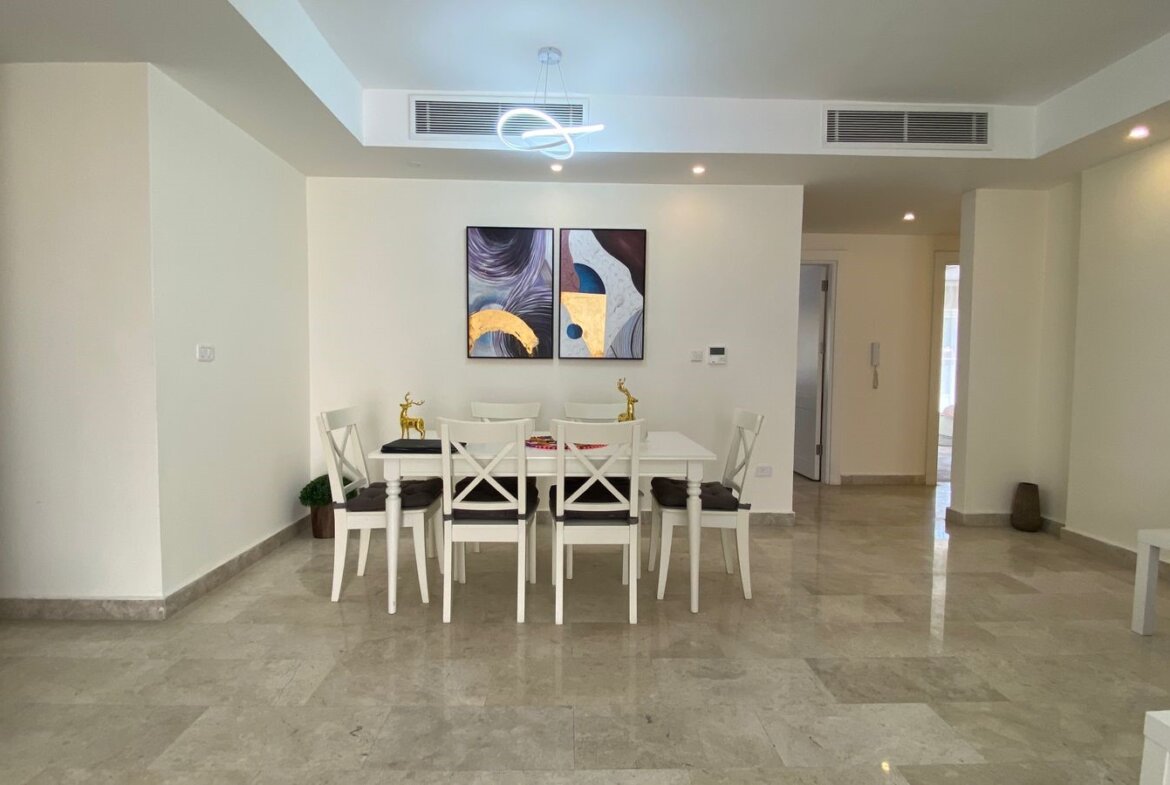 Bright dining area with a white table and six white chairs, two-panel abstract art, and gold deer figurines under a modern loop chandelier.