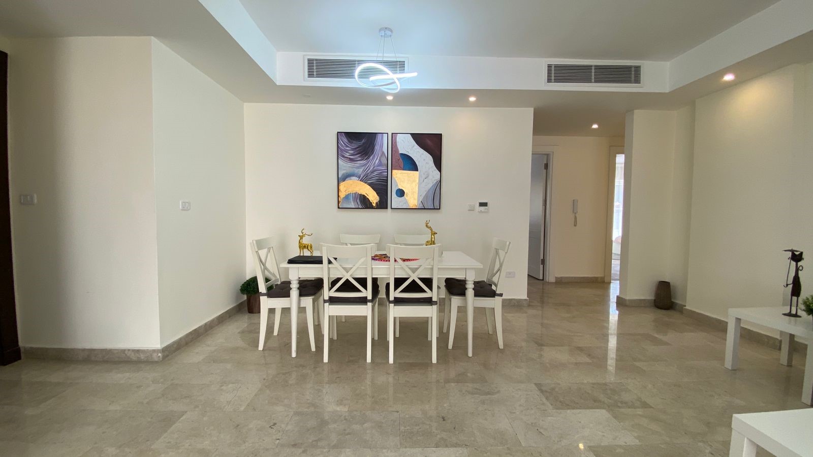 7e1f3ffc-4c5d-4513-b625-19cc381a8a08 Bright dining area with a white table and six white chairs, two-panel abstract art, and gold deer figurines under a modern loop chandelier.