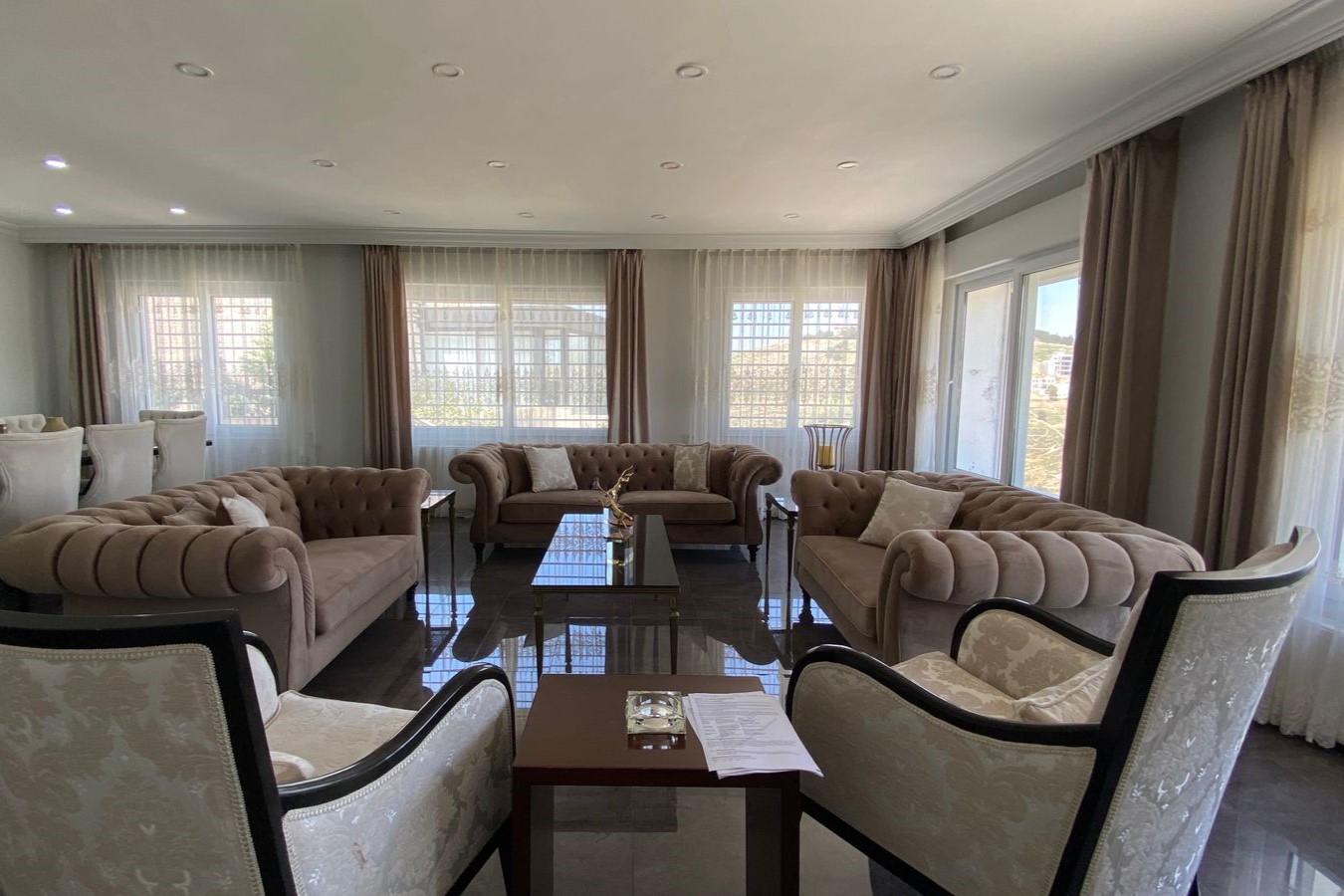 7e56bd8a-43ff-4430-ba72-f95dc6b85282 Elegant living room with tufted beige sofas around a glass coffee table, large windows with sheer curtains on both sides.