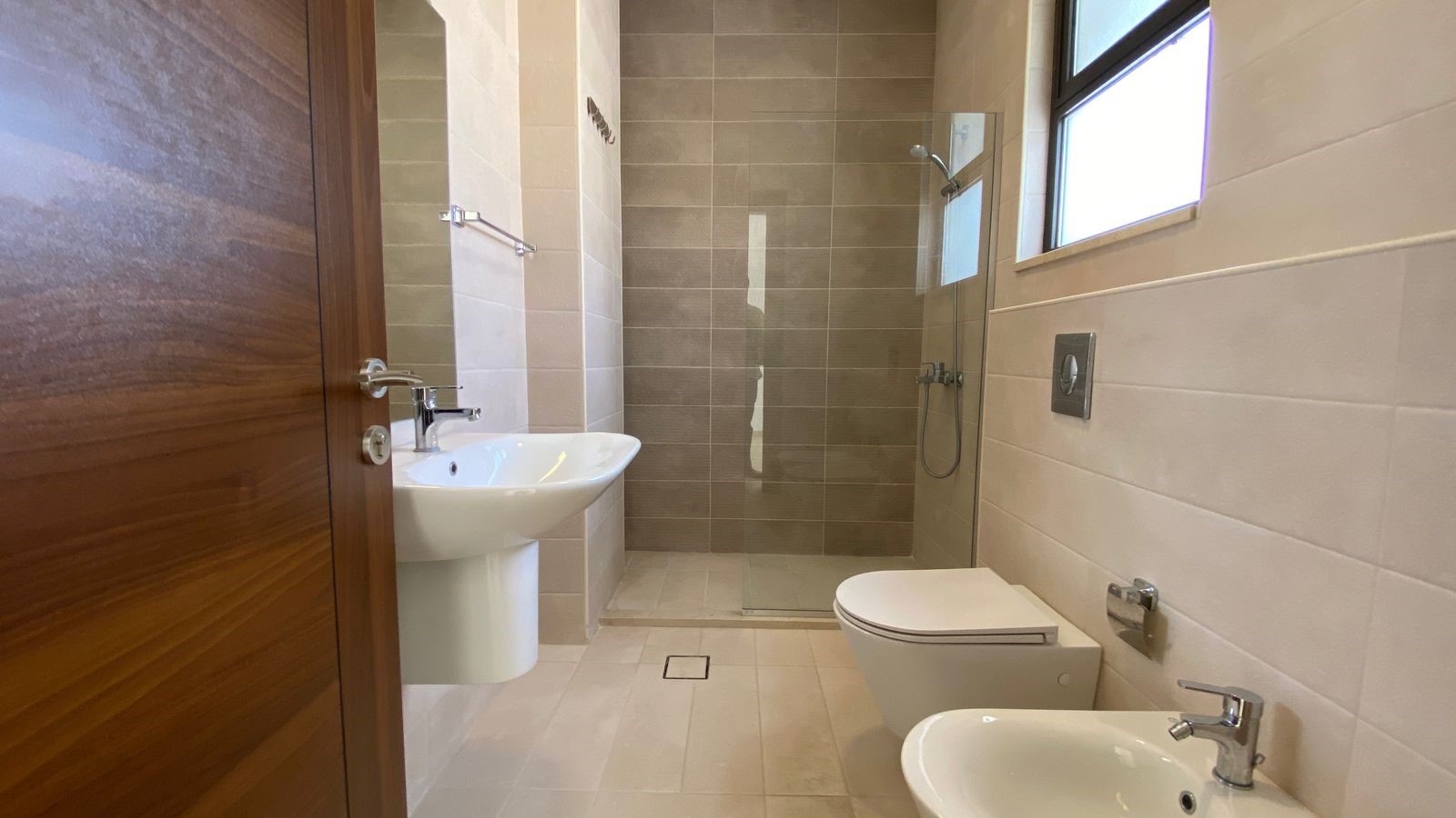 7f5240e5-640f-4e92-b301-6e0a9b6a20d2 Modern bathroom with a wall-mounted sink, glass-enclosed shower, toilet and bidet, and beige tiling far wall. A wooden door is visible on the left.