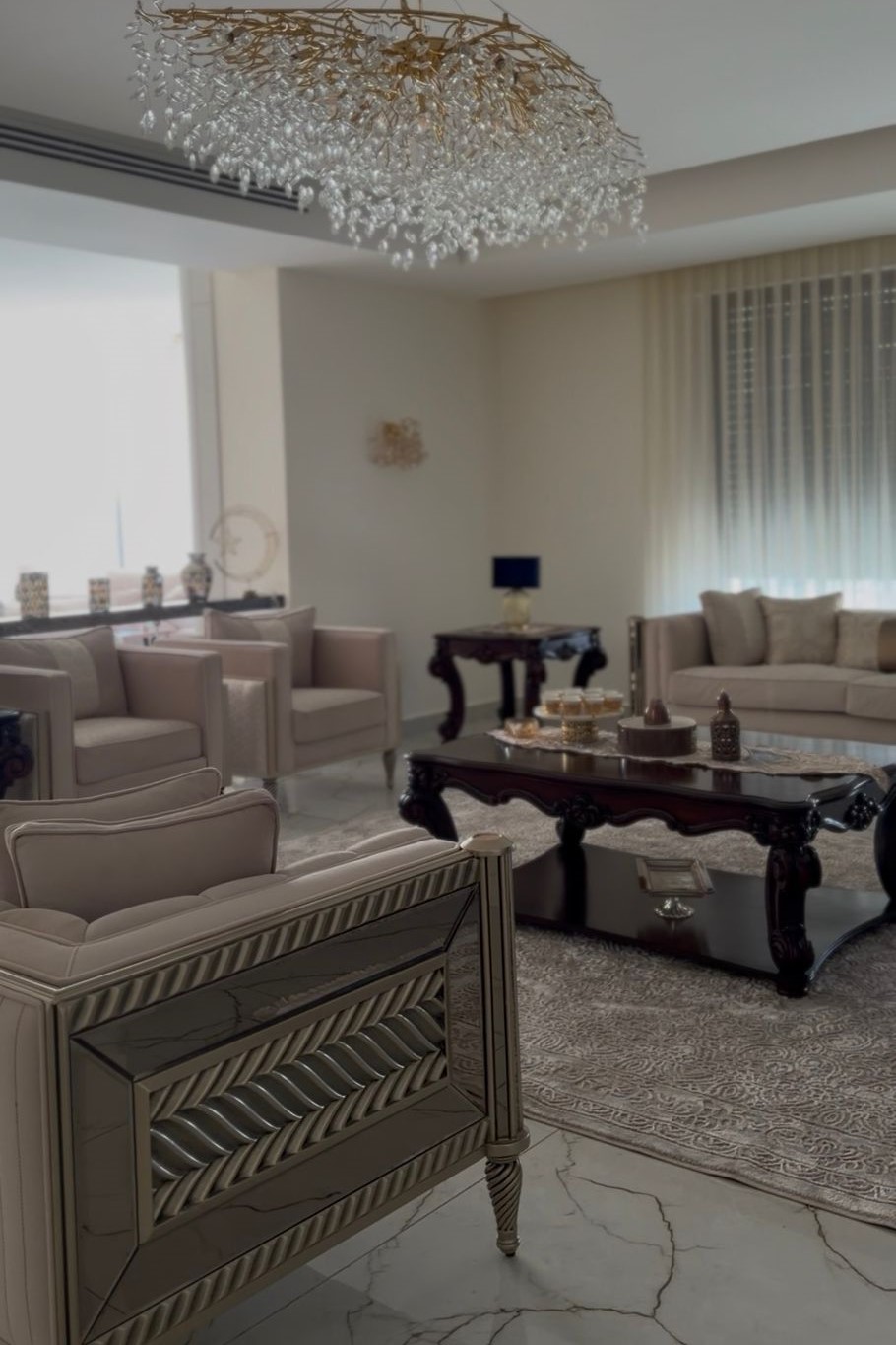 7f5c268b-4917-4c29-8bce-b14a8c8b30a7 Luxurious living room with cream tufted seating, ornate dark wood coffee table, and a crystal chandelier.