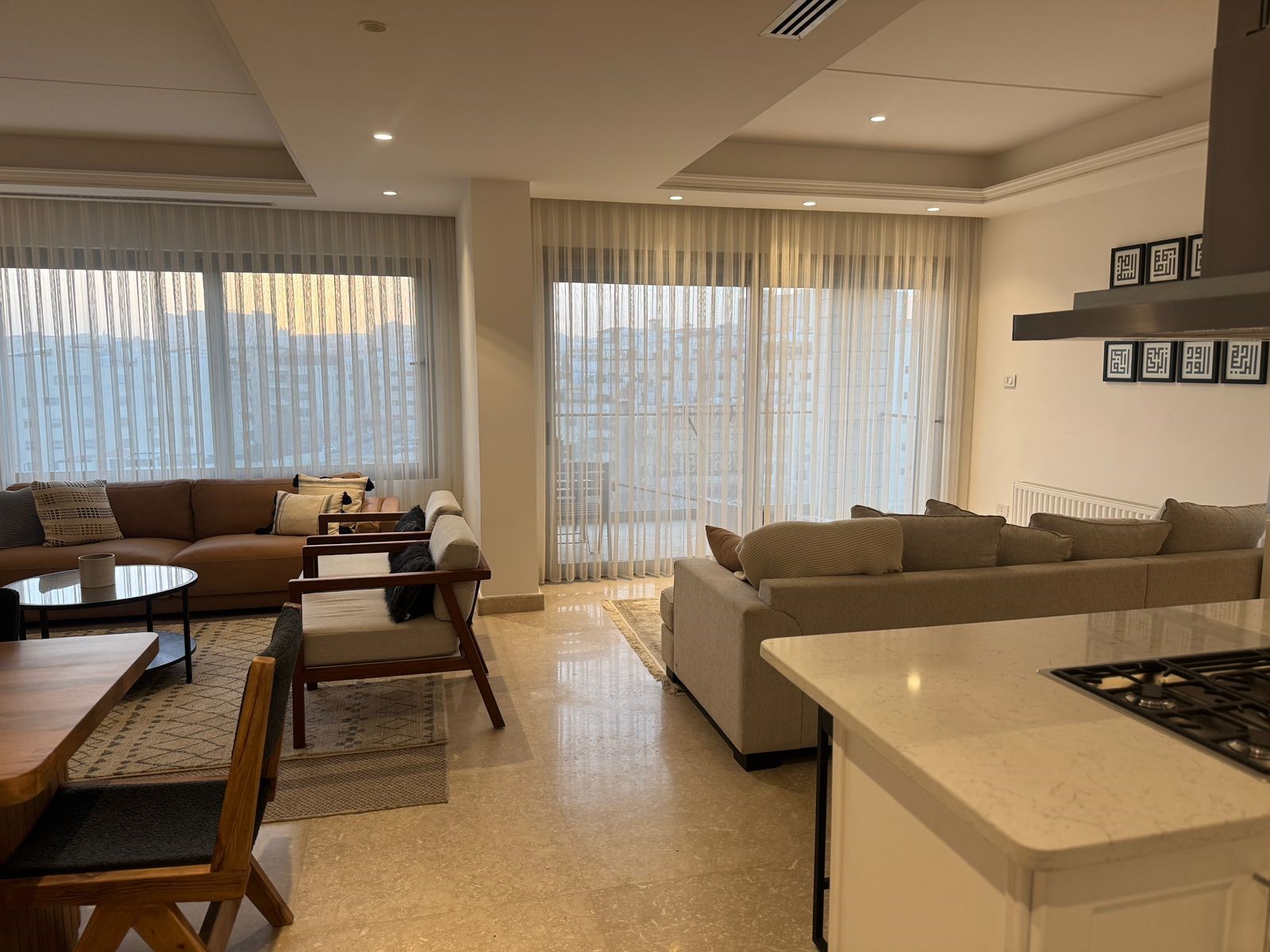 7f9d75b8-7146-4a37-b5fb-077a0859eedd Open-concept living room with beige sofas, wooden chairs, and sheer floor-to-ceiling curtains revealing a city view outside.