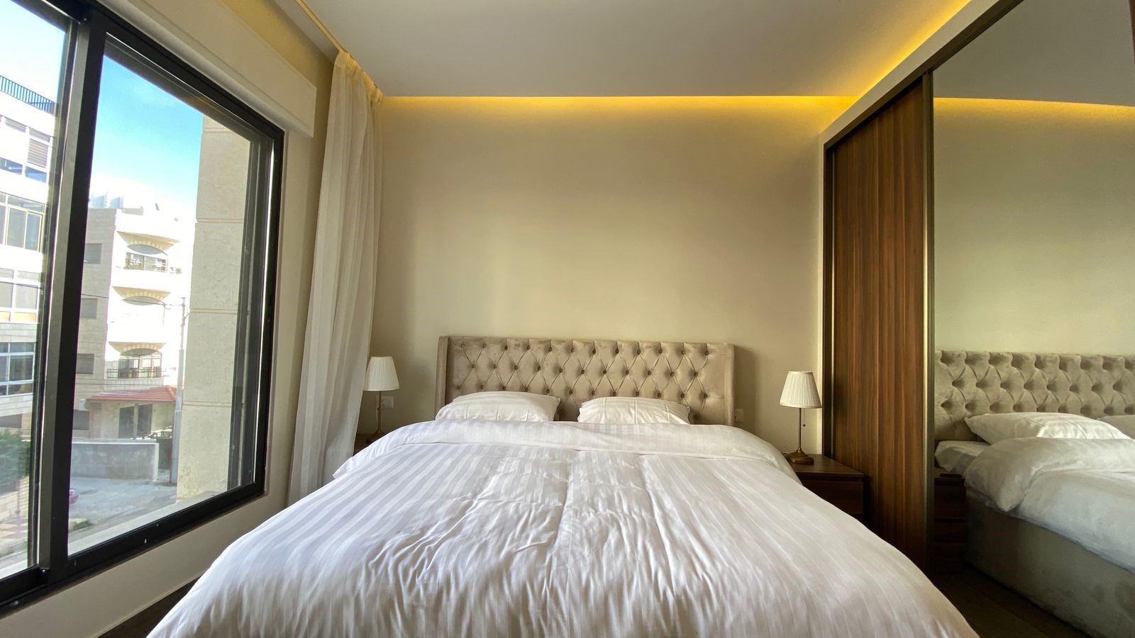 80c43399-2516-4bf2-a510-0b11ecd2d8f7 Hotel bedroom with white striped duvet, tufted beige headboard, two nightstands and lamps, and a large window to the left.