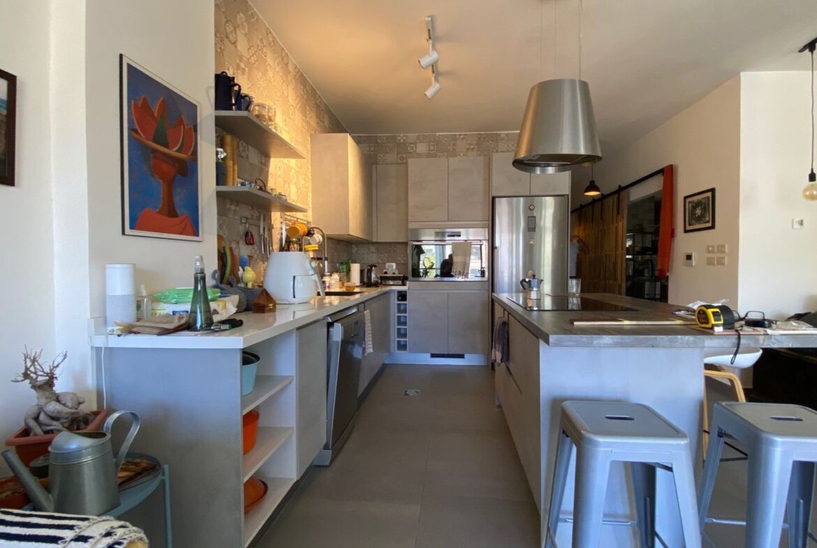 Open-concept kitchen with a long island, stainless steel appliances, and gray cabinets; metal bar stools along the island edge.