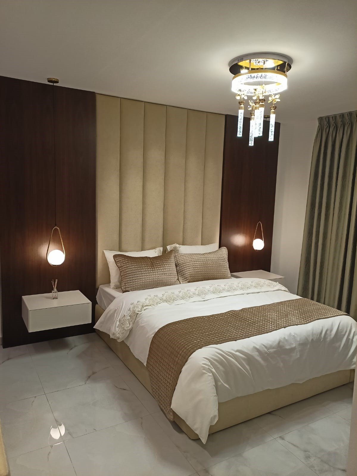 818e0bde-1149-48db-beaa-4afa2ecd79c7 Luxurious beige-brown hotel-style bedroom with a padded headboard, pendant lamps, and a chandelier above the bed.