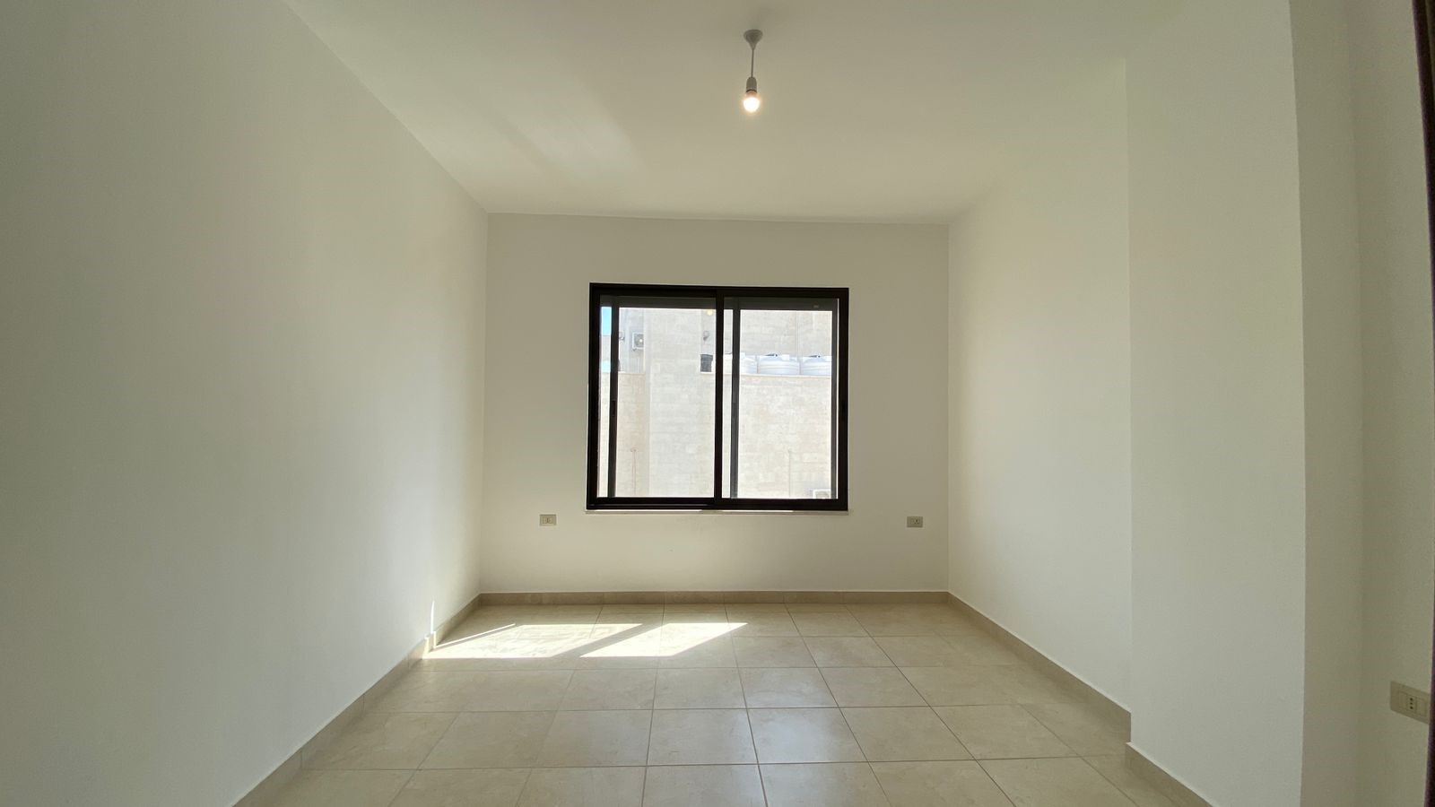 81f8eef6-2ebc-42a3-b245-b46fa03326f3 Empty rectangular room with white walls and beige tile floor, a large black-framed window letting in daylight, and a single hanging bulb at the ceiling.