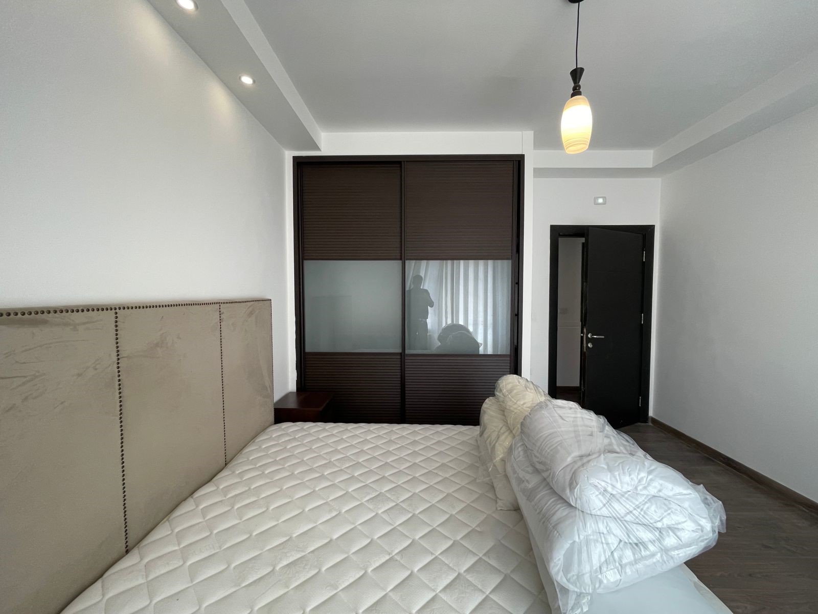 827a1e84-012c-401a-a397-704a5378dcdd Bedroom with a beige upholstered headboard, white quilted mattress, and a dark wood sliding-door wardrobe with frosted panels; pendant light hangs from the ceiling.