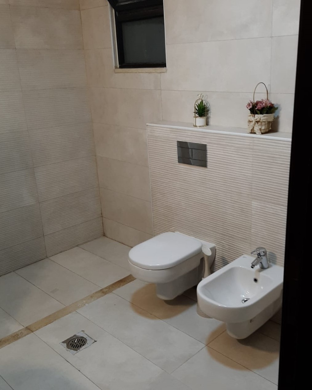 8306b236-75db-4032-a7d5-ad34e83f6d33 Modern beige-tiled bathroom with a wall-mounted toilet and a separate bidet, beige floor tiles, and a small window above. There are decorative plants on a ledge behind the fixtures.