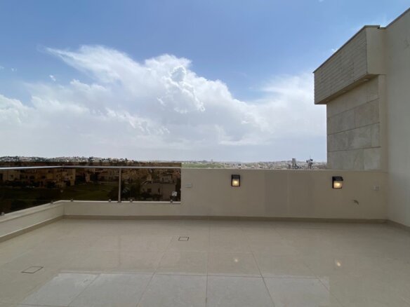 Wide rooftop terrace with beige tile flooring, glass railing, and a distant cityscape under a blue sky with clouds.