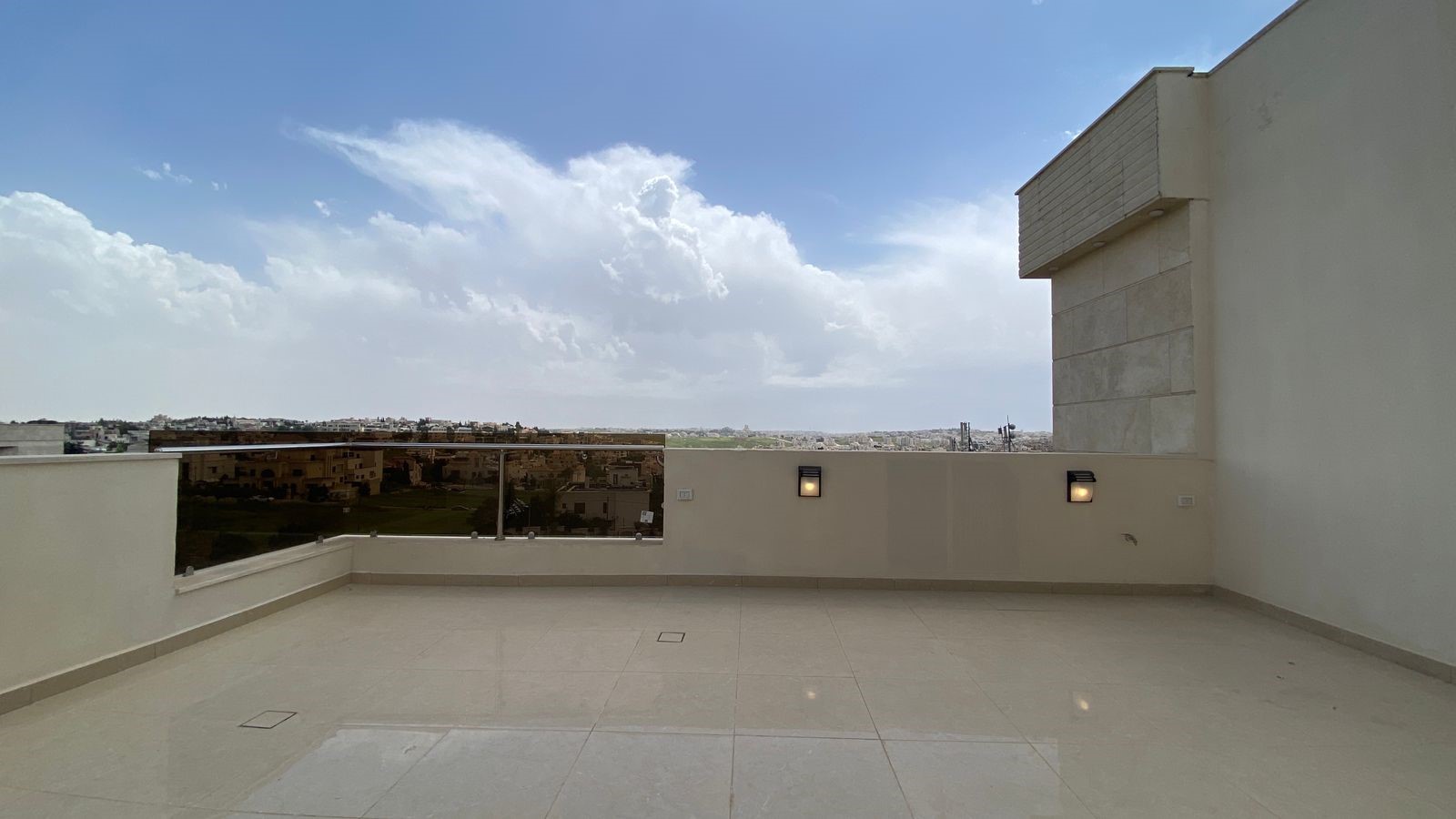 841b1ed3-19d7-4df0-9ba8-40d7a18842b5 Wide rooftop terrace with beige tile flooring, glass railing, and a distant cityscape under a blue sky with clouds.