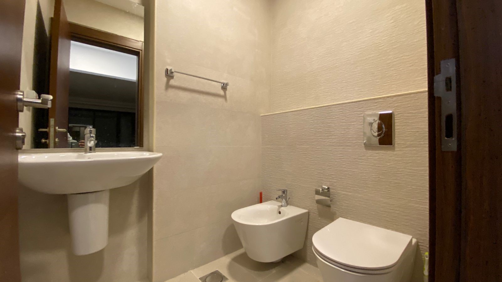 847e1c12-ae57-4645-b380-77f4026d0ec7 Compact beige-tiled bathroom with a white pedestal sink, mirror, bidet, and wall-mounted toilet side by side.