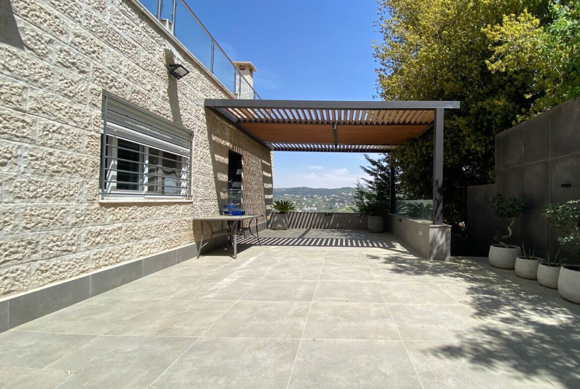 Spacious tiled rooftop terrace with a wooden slatted pergola, stone exterior wall, and view of the city beyond, with potted plants along the edge.