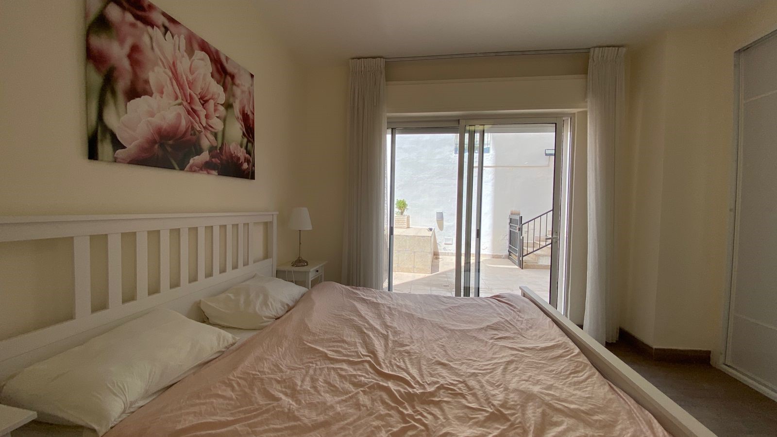 85e67955-8700-47e7-b2d8-5003d915a166 Bedroom with a white wooden bed, pale pink duvet, and two white pillows, a small nightstand with a lamp, and a large sliding glass door that opens to a sunlit balcony with outdoor stairs and railing; above the headboard hangs a framed pink floral canvas.
