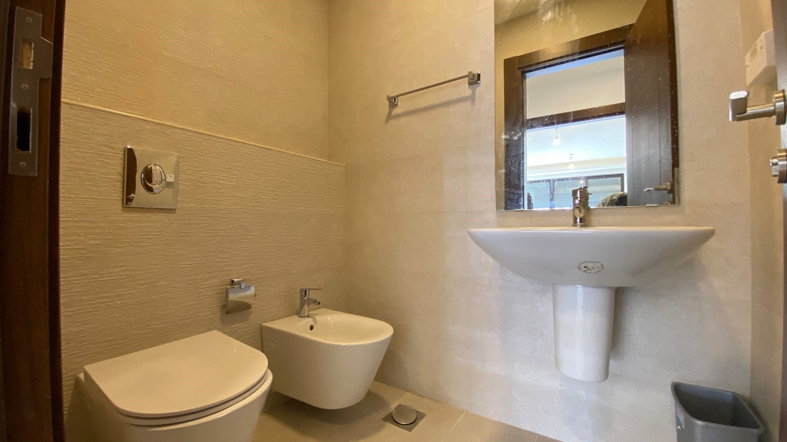 86422607-d8a0-4809-877e-7c2d2d37d9ee Modern beige-tiled bathroom with wall-mounted toilet and bidet, pedestal sink, and rectangular mirror above the sink viewed from doorway.