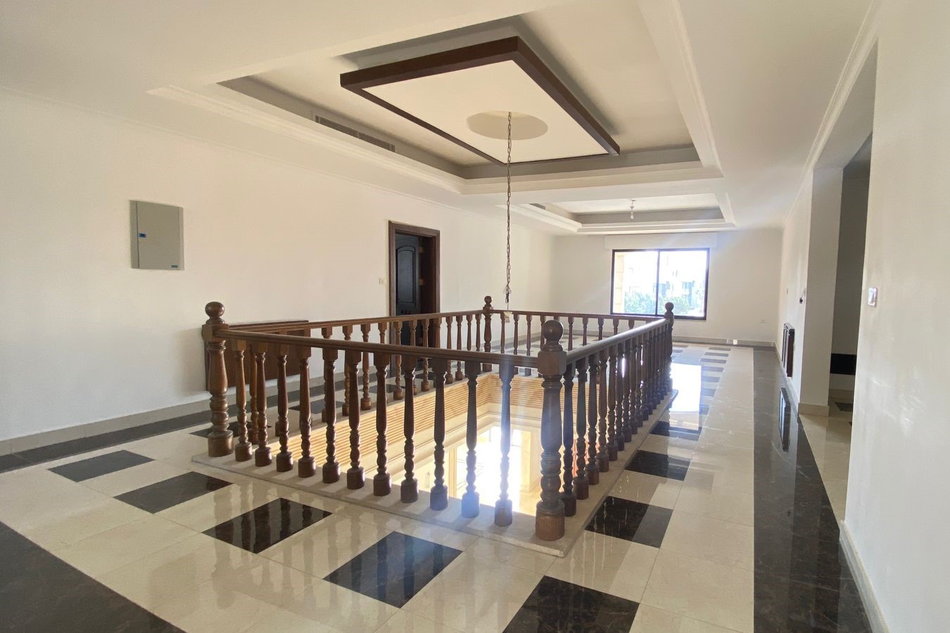 868806bb-55a3-4814-886d-1ae1ad186da9 Spacious hallway with a wooden balustrade surrounding a square opening, black-and-white checkered marble floor, and a coffered ceiling.