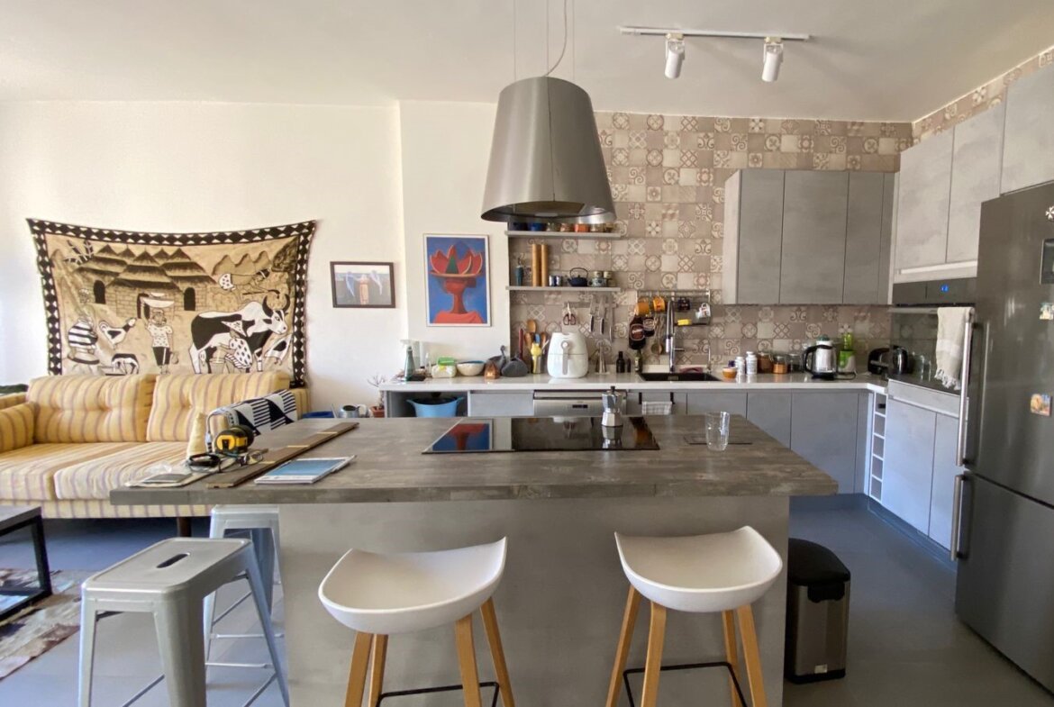 Open-plan kitchen with grey cabinets, patterned tile backsplash, and a large central island; two white stools sit at the island edge.