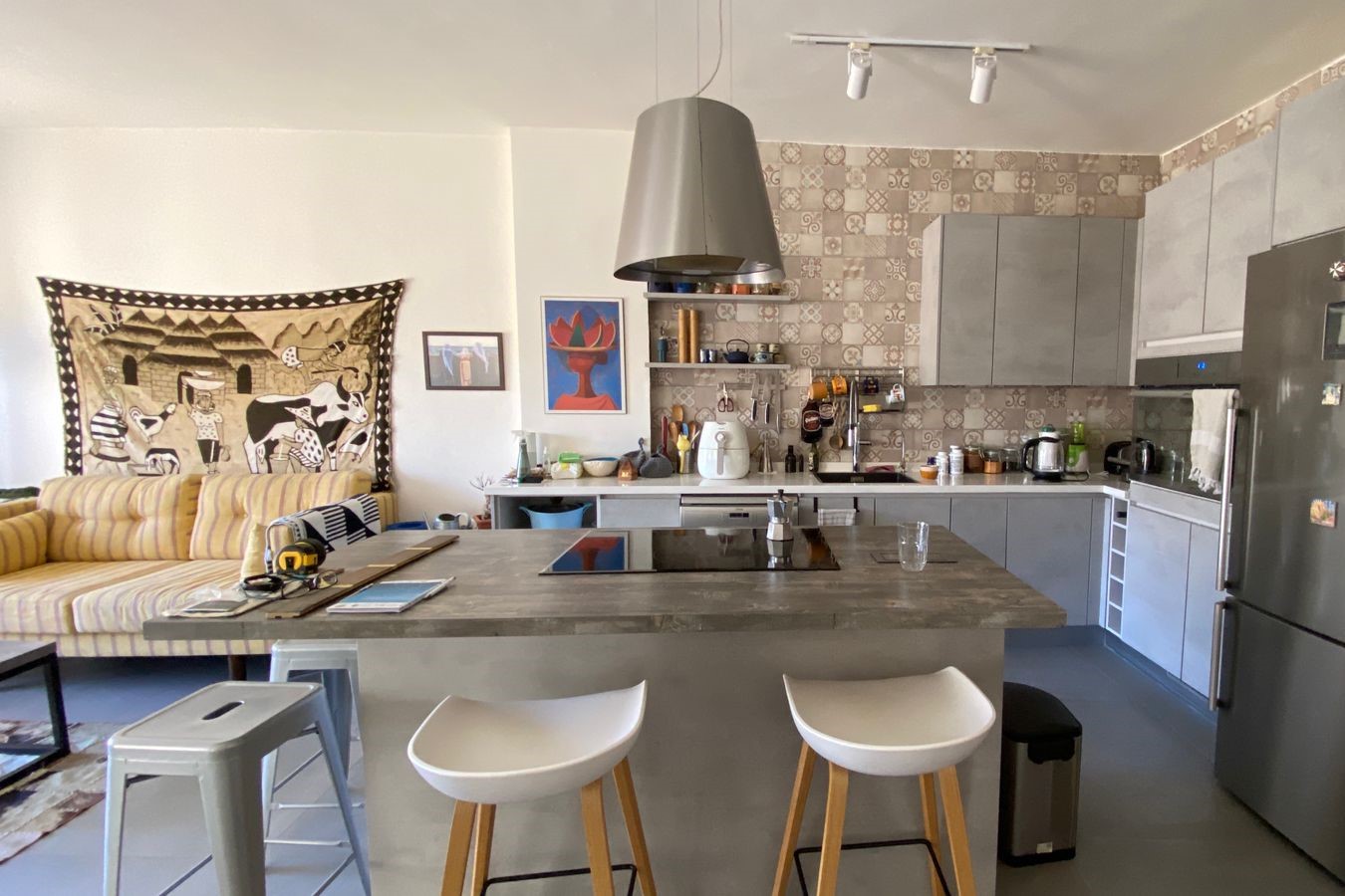 Open-plan kitchen with grey cabinets, patterned tile backsplash, and a large central island; two white stools sit at the island edge.
