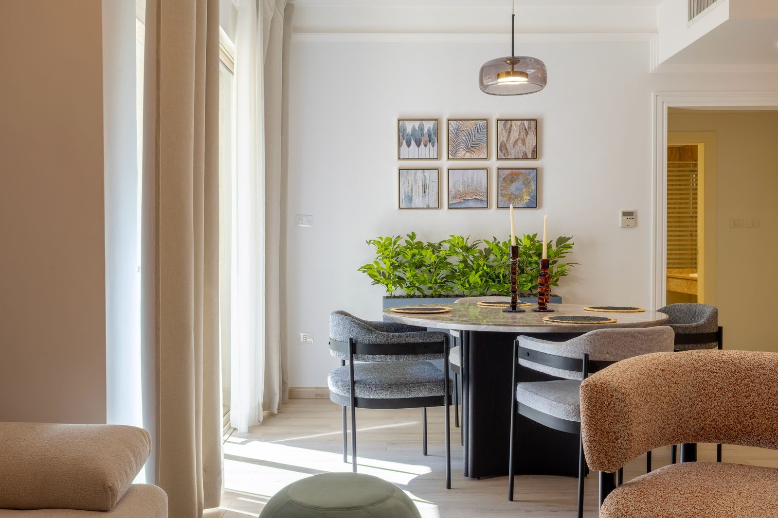885a75eb-2552-41c7-81c5-0c19dbf019ff Bright dining area with a round marble table, six gray upholstered chairs, and a row of six framed botanical prints on a white wall beneath a pendant light.
