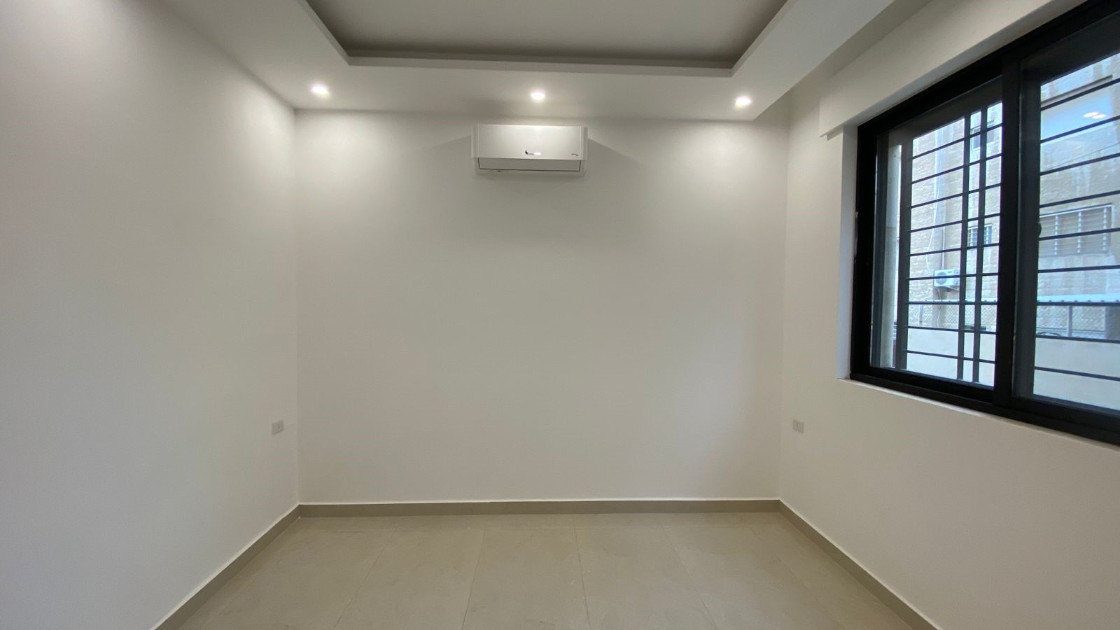 8892bddf-86f9-4161-b974-953619db1503 Empty white room with tiled floor, wall-mounted AC, recessed ceiling lights, and a large barred window.