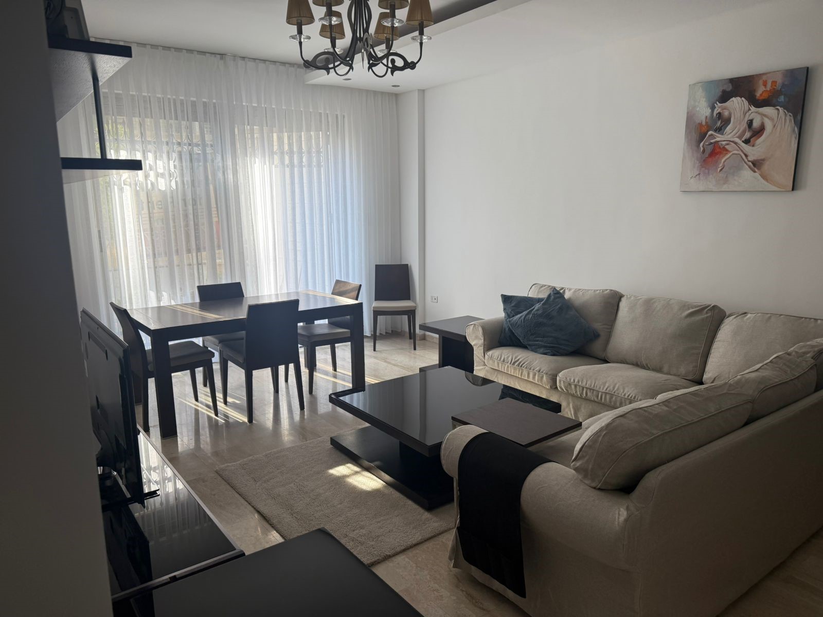 88a650b0-15eb-4e2d-b1ab-81c052f8a33c Bright living and dining area with a beige sofa, black coffee table, and a dark wood dining table set by large windows with sheer curtains.
