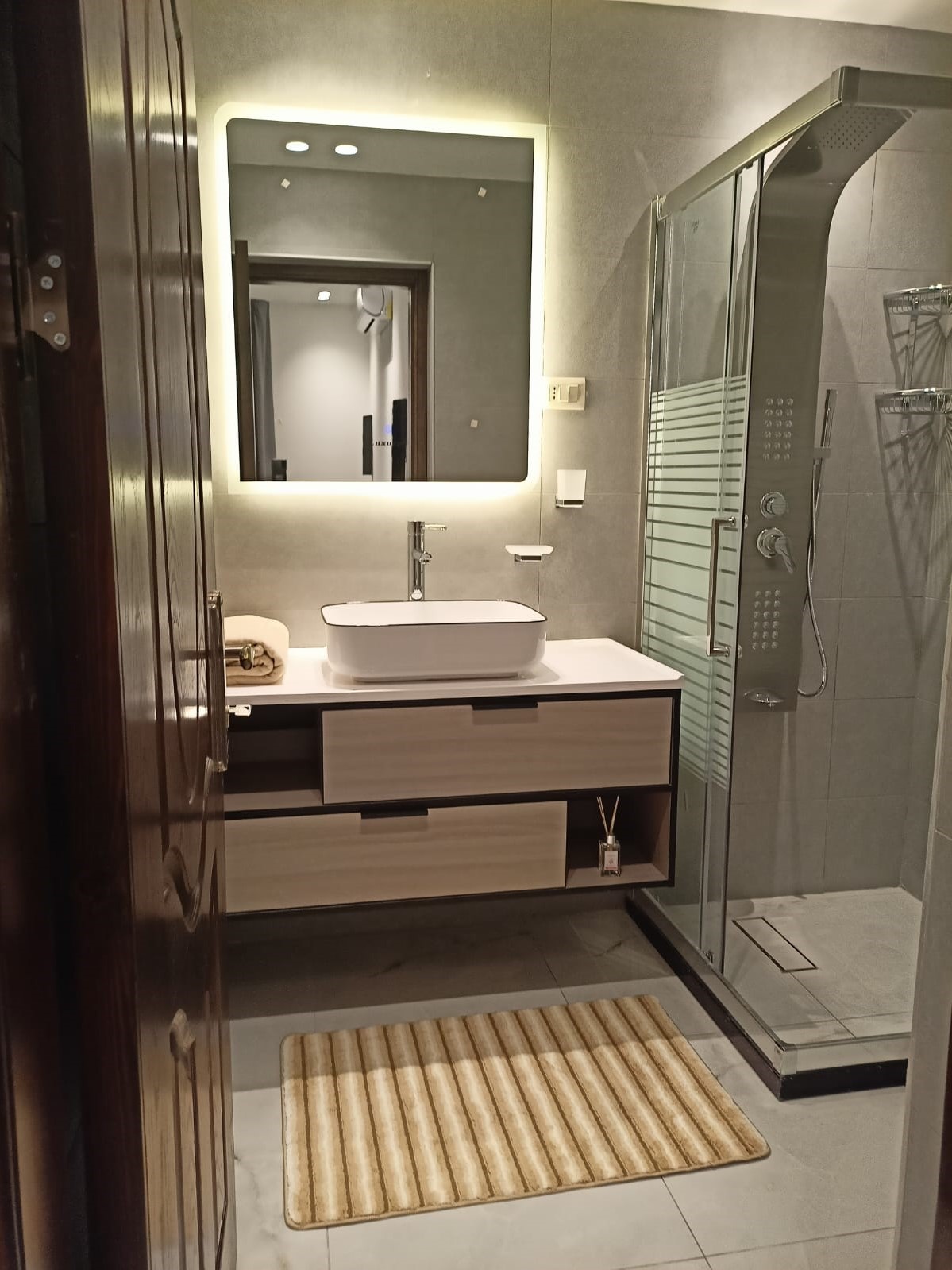 88d78b98-6e1d-47aa-8ef6-7e9937e19062 Modern bathroom with a backlit rectangular mirror above a white vessel sink on a floating wooden vanity, beige striped bath mats on tiled floor, and a glass shower enclosure to the right.