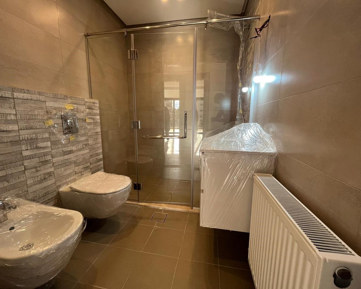 8a111491-9ad9-4c48-b84a-ef6be4b9e017 Modern bathroom under protection: toilet, bidet, sink, and shower enclosure all wrapped in plastic during construction or renovation, with a radiator along the wall.