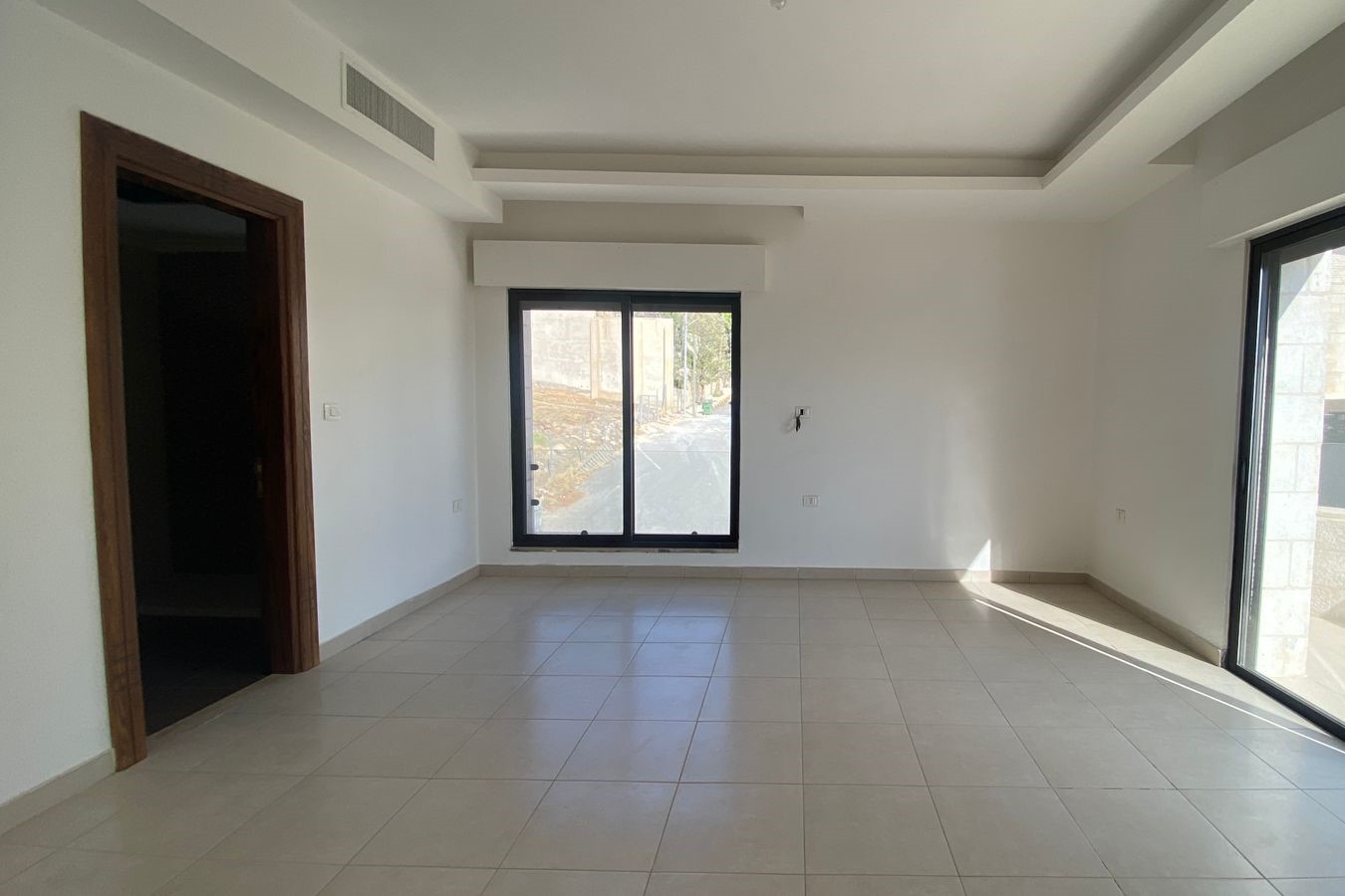 8a566d08-6d0e-41f0-9809-d7e51b236847 Unfurnished living room with large sliding glass door to outside, white walls, beige tiled floor, and a wooden doorway on the left.
