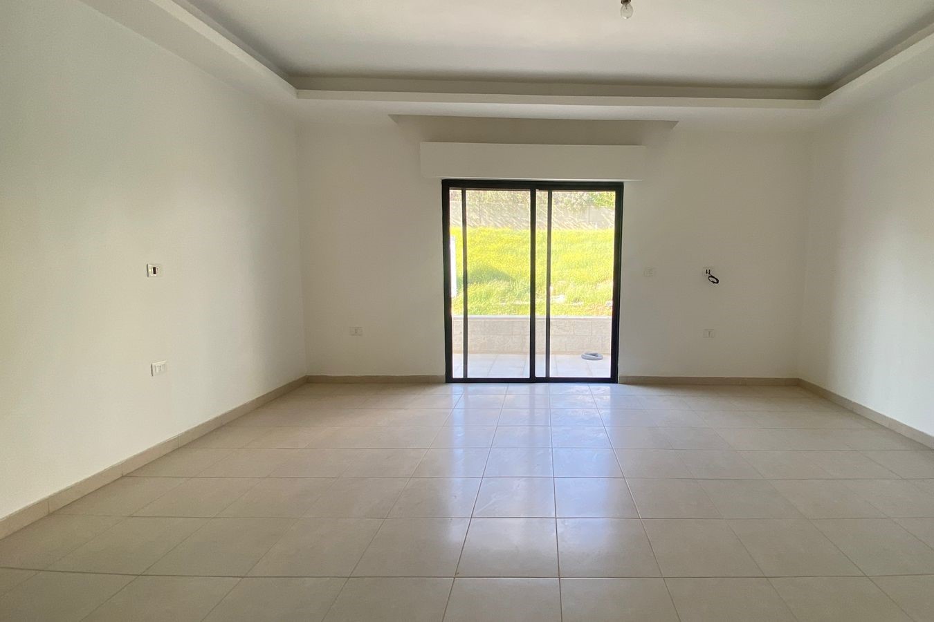 8a68591f-3891-4c49-8d9e-d28f77b2449a Empty living room with beige tile floor, white walls, and a sliding glass door to the patio/outdoor area.