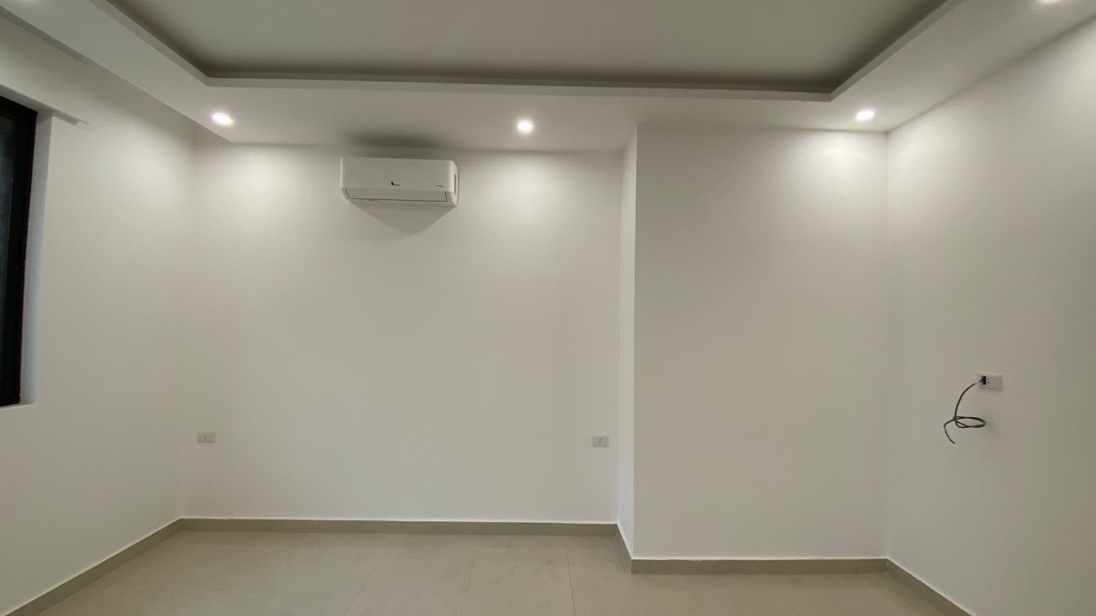 8ad3a245-c0ed-4b10-b5e3-cd370041a30c Empty white room with beige tiled floor, wall-mounted AC, recessed ceiling lights, and a window on the left.