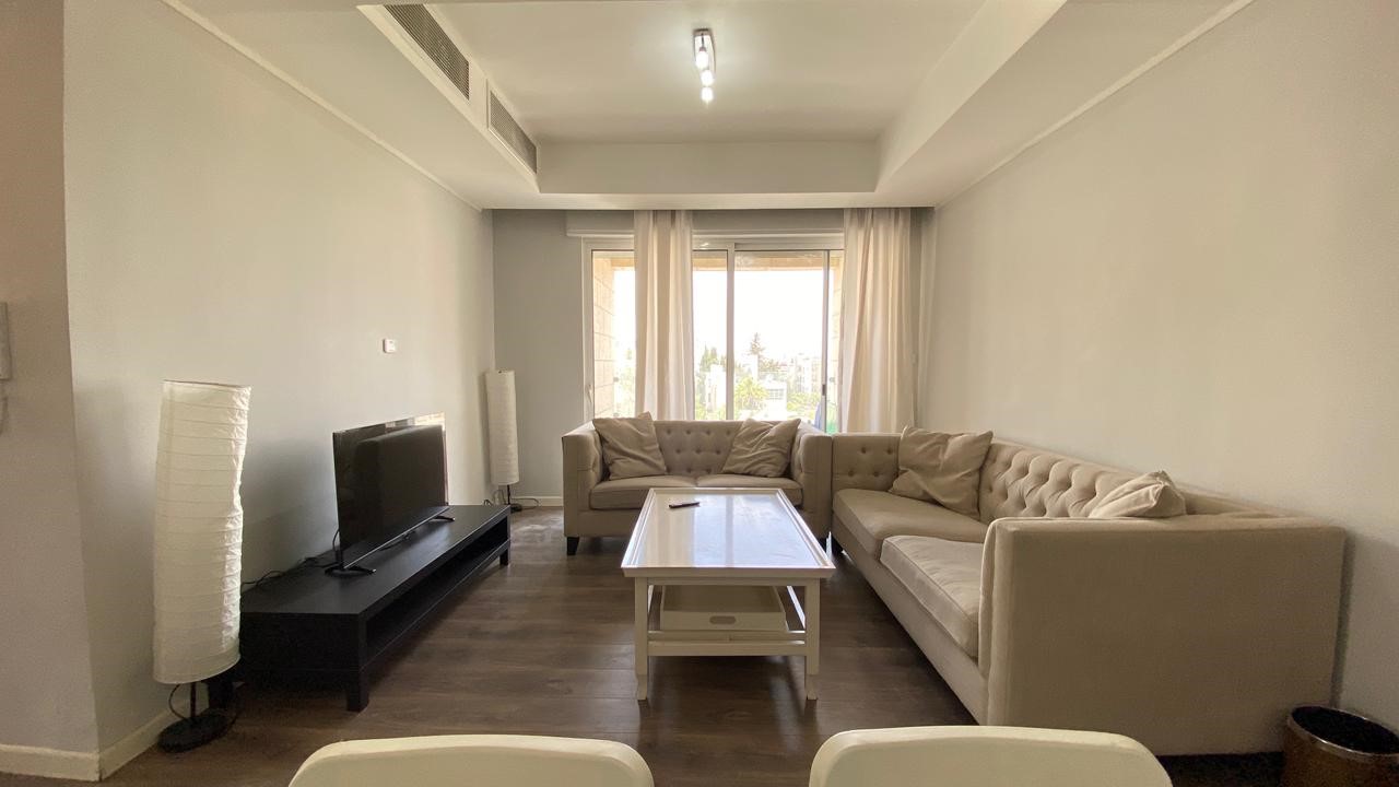 8c7168c2-972c-4417-b46f-11a200ffc3e3 Living room with a beige sectional sofa, white coffee table, and a TV on a black console facing the seating area.
