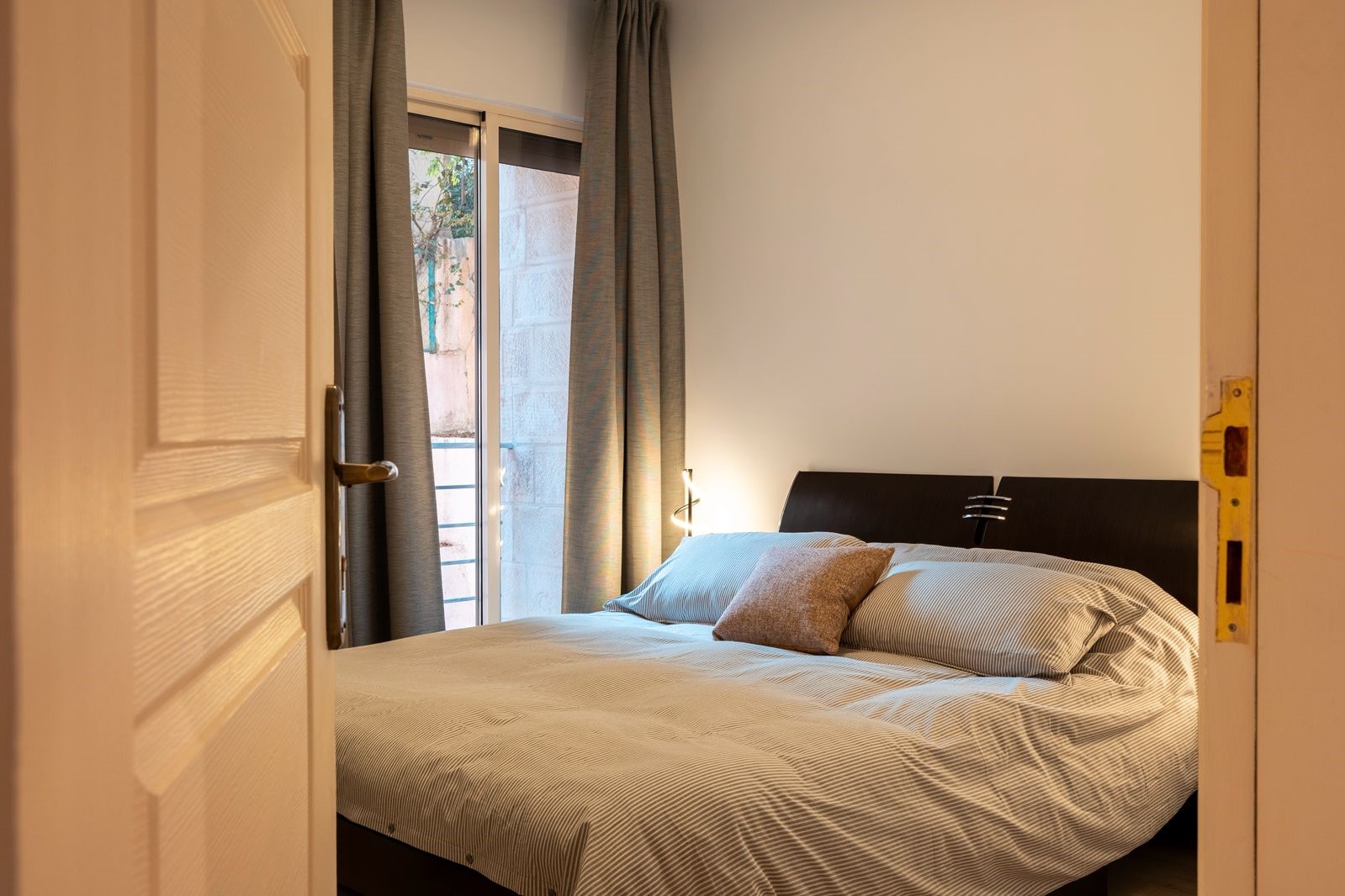 8d6c9972-861f-4f05-b013-f51546cd0d33 Bedroom with a double bed, striped duvet and brown decorative pillow, dark headboard, beside a window with gray curtains.