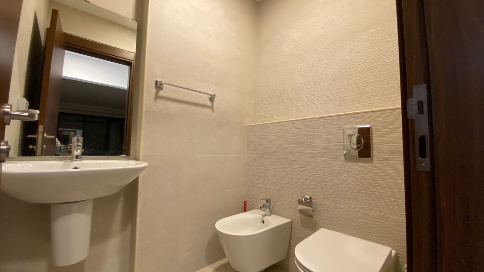 8e1af424-33f5-4f3a-9194-efafdcdb9552 Small modern bathroom with pedestal sink, mirror, bidet, and wall-hung toilet; beige tiled walls the fixtures create a compact layout.