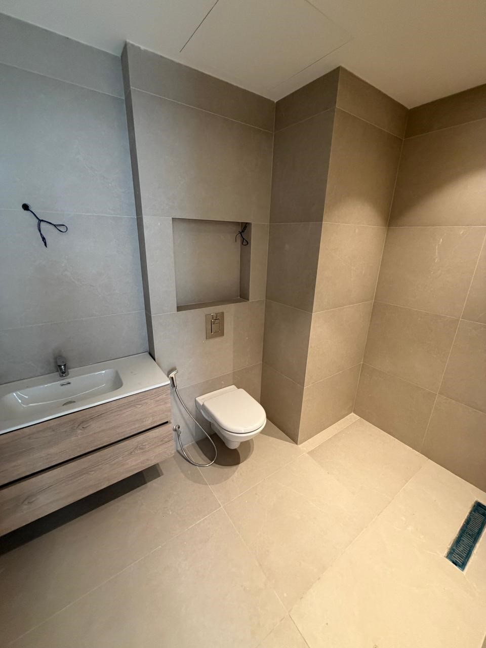 8ebb8e7e-22e9-4e59-a329-685ab8e995d0 Modern bathroom with a wall-mounted toilet, a floating wooden vanity with sink, and a recessed wall niche, all in gray tiles; a handheld bidet hose beside the toilet.
