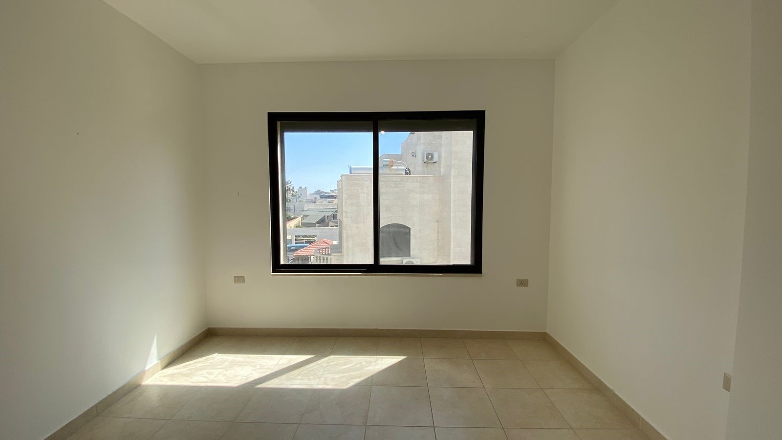 8ed7210a-ca5f-4d92-8e5f-f0f298f7bf11 Empty white-walled room with a large black-framed window; sunlight pours onto the tiled floor, urban buildings visible outside.