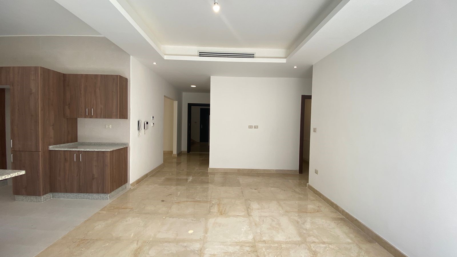 8fec63e4-dc65-4b49-bc27-b88db6b02f2b Empty open-concept living area with wood kitchen cabinets on the left, beige marble floor, white walls, and a hallway at the far end.